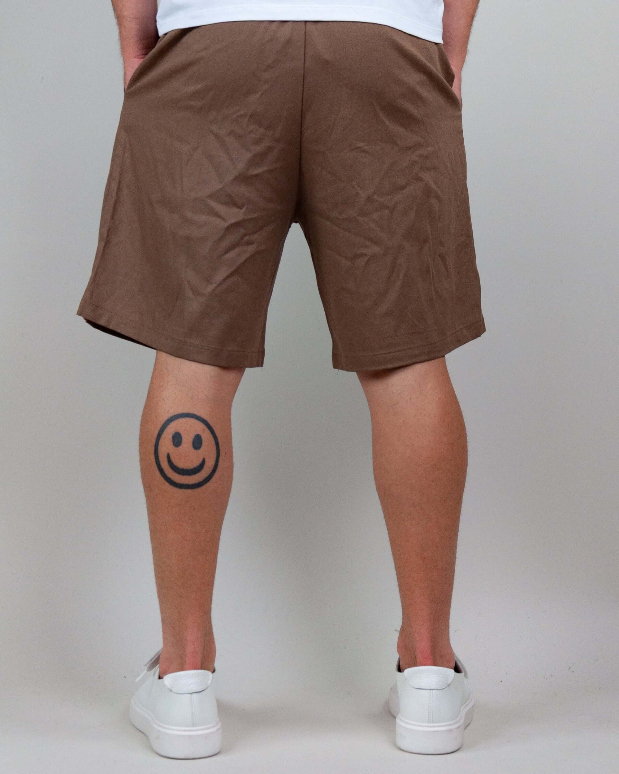 Back view of a person wearing brown Bermuda shorts and white sneakers, showcasing a smiley face tattoo on the calf.