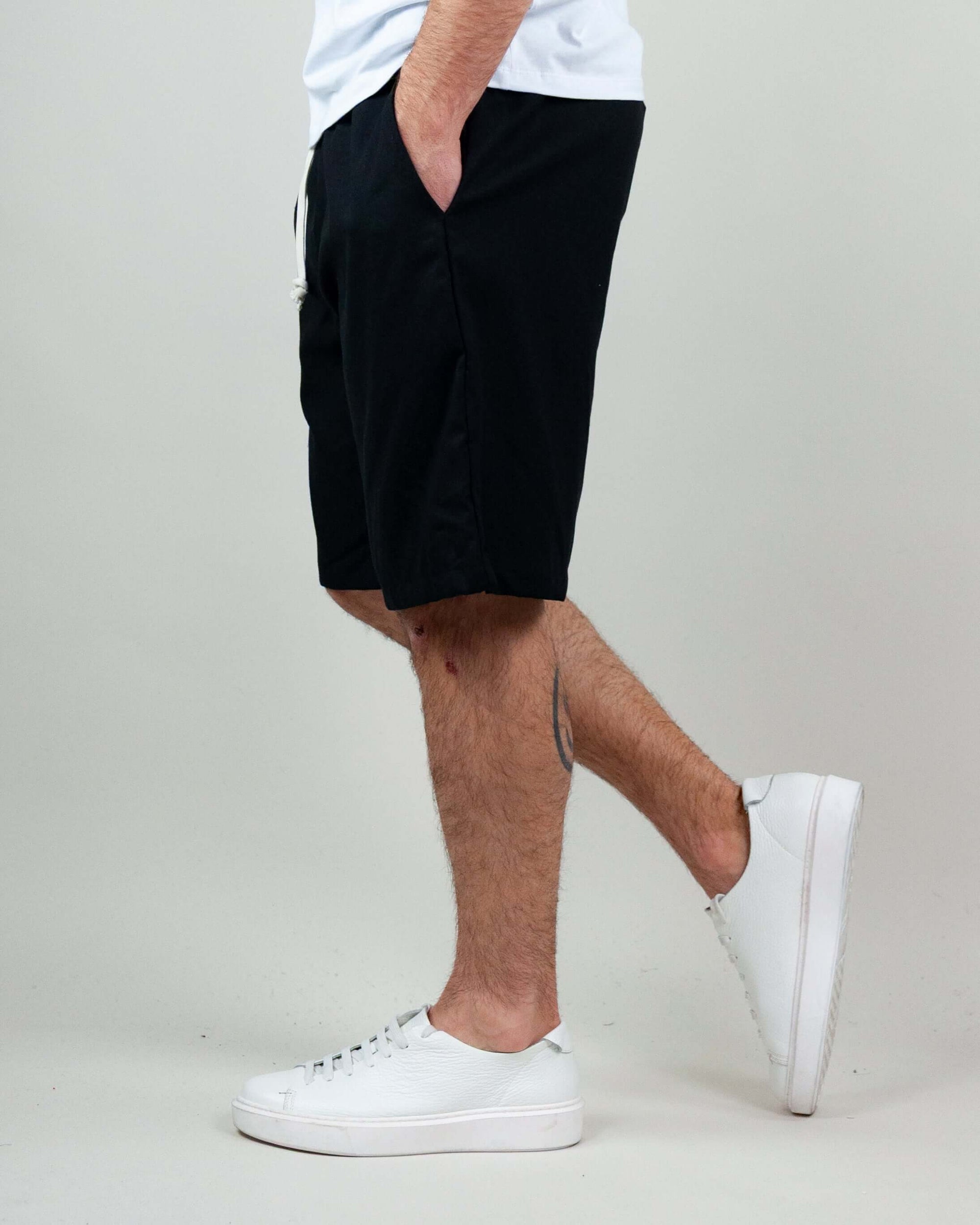 Side view of a man wearing black Bermuda shorts and white sneakers against a neutral background.