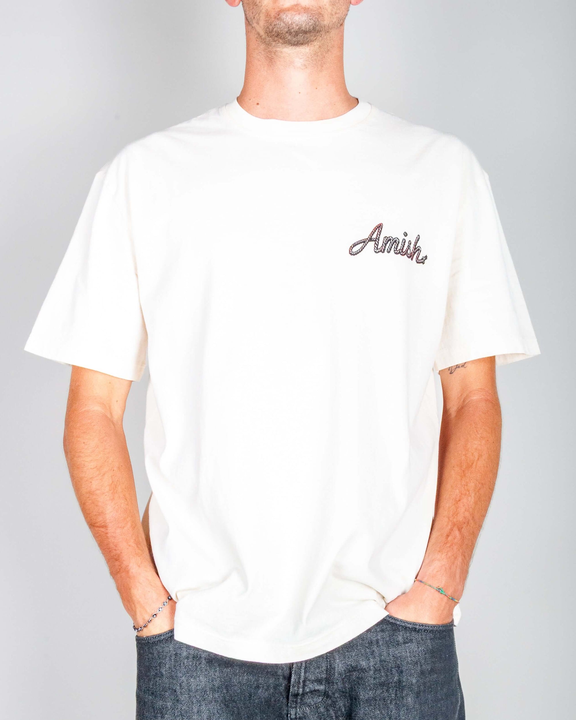 T-shirt arte cowgirl Amish color panna con logo ricamato, indossata da un uomo con mani in tasca.